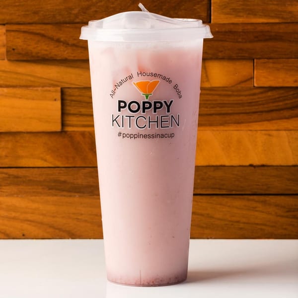 A tall plastic cup of pink boba tea with the Poppy Kitchen logo against a wooden background.