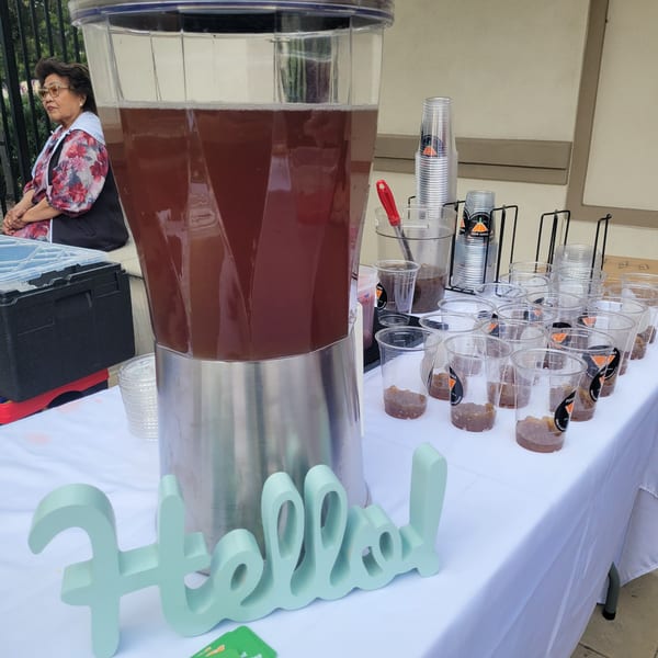 Large drink dispenser and many small cups with boba on a white-covered table, with a woman seated nearby.