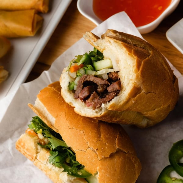 Two Vietnamese banh mi sandwiches with meat and vegetables, with spring rolls and sauce in the background.