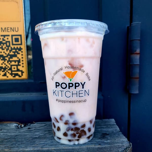 A Poppy Kitchen boba tea with a light pink drink and dark boba pearls sits on a wooden surface.