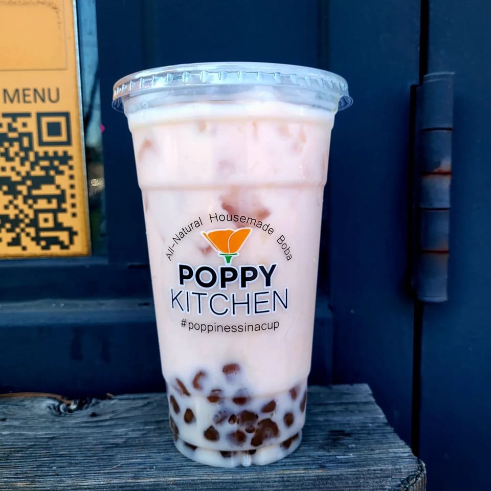 A Poppy Kitchen boba tea with a light pink drink and dark boba pearls sits on a wooden surface.
