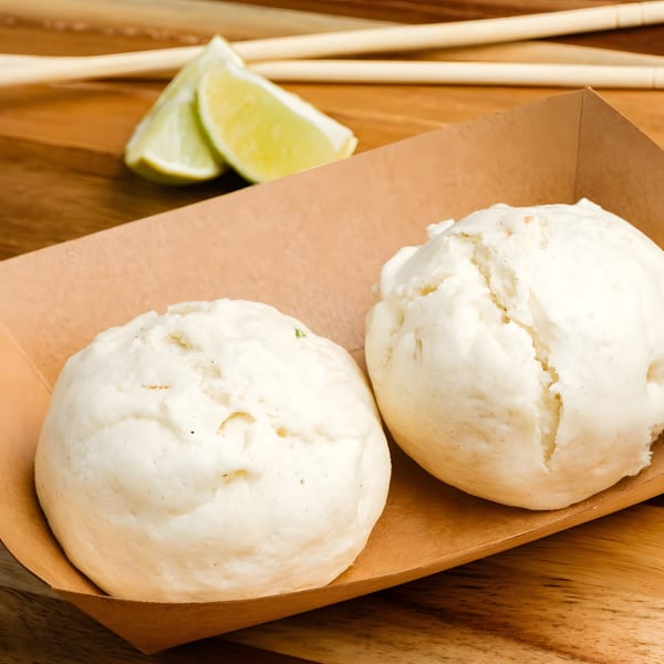 Two fluffy white steamed buns in a brown paper tray with lime wedges and chopsticks.